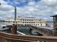 A large riverboat with multiple decks is docked by a wooden pier on a cloudy day.
