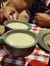 Two people are clinking mugs filled with creamy soup over a checkered tablecloth.
