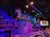 The image depicts a theater stage set resembling a rustic, multi-level treehouse with colorful lighting, surrounded by an audience seated at tables.