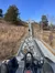 A person is riding a roller coaster or alpine coaster up a hill on a sunny day, with trees and dry grass on either side.