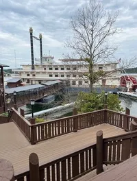 A large riverboat with two tall smokestacks is docked beside a wooden walkway and stairway under a cloudy sky.