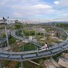 A person is riding a twisting alpine coaster with a scenic view of a town and mountains in the background.