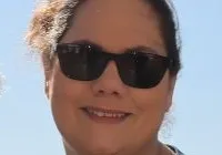 A man and a woman are standing beside a ship's wheel on a sunny day, with water and a railing in the background.