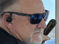 A person wearing sunglasses and a headset stands at the helm of a boat, looking out over a scenic body of water through large windows.