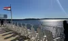 The image shows a row of white Adirondack chairs on a sunlit deck overlooking a sparkling body of water, with a flag waving and a few people enjoying the view.