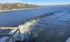 A paddlewheel boat is moving through a wide river, leaving a wake in the water, with a tree-lined shore in the background under a clear blue sky.