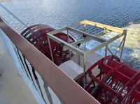 A red paddlewheel on a riverboat is churning through water creating a wake.