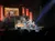 A band is performing on stage with a backdrop featuring rock posters and a large image of The Eagles' album cover.