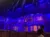 The image depicts a stage set with an ornate architectural design, illuminated in blue and purple lights, with people visible in the foreground.