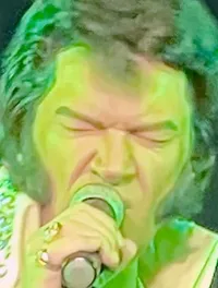 A performer dressed in a rhinestone-studded white jumpsuit sings into a microphone on stage under green lighting.
