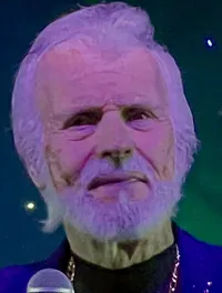 A man with a beard, wearing a blue jacket and holding a microphone, stands on a stage with a starry backdrop.