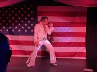 A performer dressed in an Elvis-inspired outfit sings in front of a large American flag backdrop.