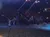 The image shows a dimly lit indoor arena with a performance involving camels and riders, while an audience watches from tiered seating.