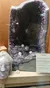 The image shows a large amethyst geode on display with a necklace and an informational plaque nearby, along with a carved head sculpture.