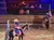Two people on horseback, wearing patriotic outfits and holding American flags, perform in an indoor arena with spectators watching from an elevated seating area.