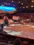 The image captures a Wild West-themed stage performance featuring covered wagons, horses, and performers in a large arena with an audience seated around the edges.