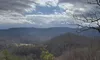 The image depicts a scenic view of a mountainous landscape under a partly cloudy sky, with a leaf-strewn dirt path in the foreground.