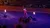 A performer in a red costume stands balancing on two horses, waving to an audience in an arena with flaming torches.