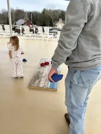 A person in jeans and a gray sweatshirt holds a blue beanbag, preparing to play cornhole on a deck, while a child stands nearby.