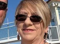 A couple wearing sunglasses takes a selfie on a boat with smokestacks and festive decorations in the background.