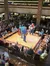A band is performing on a central wooden stage surrounded by a seated audience in a multi-level venue.