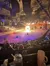 A live performance is taking place in an indoor arena, featuring rows of seated spectators and a large ring of fire at the center of the stage.