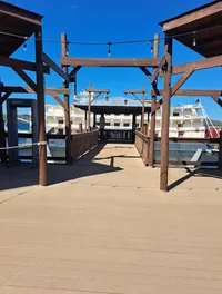 A wooden walkway with overhead string lights overlooks a partially visible riverboat on a bright, clear day.