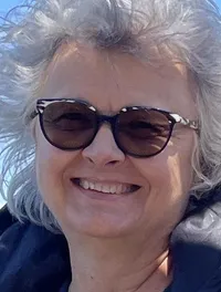 A person wearing sunglasses and a jacket stands on a balcony overlooking a body of water, holding a hat and smiling under a clear blue sky.