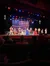 Performers in patriotic red, white, and blue costumes sing on stage in front of a large American flag backdrop.