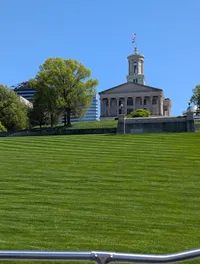 A historic building with columns and a cupola is positioned atop a grassy hill, flanked by trees and modern buildings under a clear blue sky.