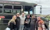 A group of six people is posing happily in front of a large green bus under a cloudy sky.