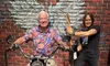 An older man is smiling while sitting on a motorcycle, with a brick wall featuring wings painted behind him and another person standing beside with a guitar.