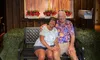 A smiling couple sits on a bench under a sign that reads 