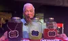 An older man playfully sticks his tongue out while posing behind three jars labeled
