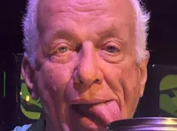 An older man playfully sticks his tongue out while posing behind three jars labeled 