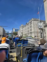 People are sitting on the upper deck of an open-top tour bus, passing a large stone building with a flag and Masonic symbol under a clear blue sky.
