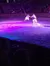 A person on horseback, galloping in an arena, holds hands with another person leaping beside them amidst colorful stage lights.
