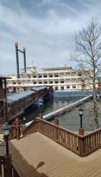 A riverboat with multiple decks is docked alongside a wooden boardwalk under a partially cloudy sky.