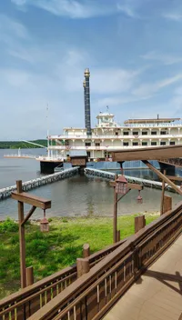 A large riverboat is moored by a wooden walkway on a calm waterfront under a blue sky.