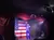 A performer in a blue jumpsuit entertains on stage with a large American flag backdrop.