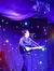 A performer is playing an acoustic guitar and singing on a stage with a starry backdrop and purple drapery.