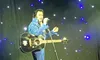 A performer in a blue, sparkling jumpsuit is singing and playing a guitar on stage.