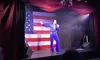 A performer in a blue jumpsuit is singing on stage in front of an American flag backdrop.