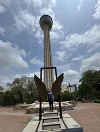 A person stands on a step in front of large metal wings, with a tall tower rising in the background beneath a partly cloudy sky.