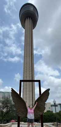 A person stands in front of a wing sculpture with their arms raised, positioned against the backdrop of a tall observation tower on a cloudy day.
