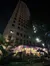 The image shows a tall building at night with a brightly lit restaurant or bar at its base, adorned with colorful string lights.