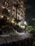 The image shows a nighttime scene near a multi-story building with balconies and palm trees, adjacent to a stone walkway and a small waterfall.