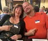A smiling man and woman sitting together at a table, each wearing name tags and pointing playfully at each other.