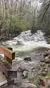 A flowing river surrounded by lush green trees and rocks, with a wooden railing in the foreground.