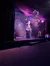 A performer in a sparkly jacket is on a dimly lit stage with decorative lights and a backdrop, gesturing towards the audience.